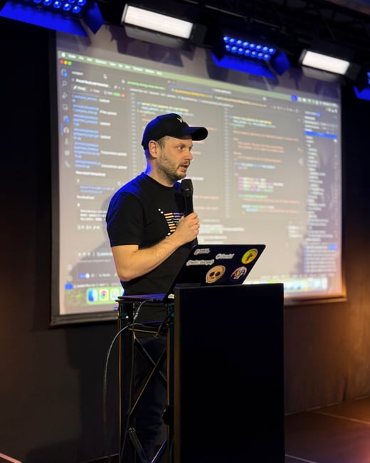A man in a black cap and t-shirt is giving a presentation on stage, holding a microphone. Behind him is a large screen displaying lines of computer code.