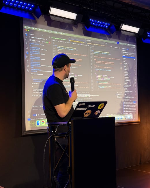 A man stands on a stage holding a microphone, giving a presentation in front of a large screen displaying Python code in an IDE.