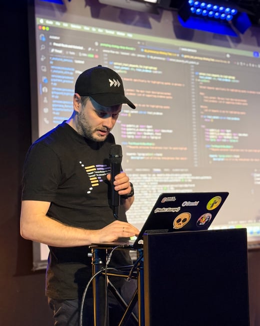A man is giving a technical presentation or live-coding demo, speaking into a microphone while using a laptop in front of a large screen displaying code.