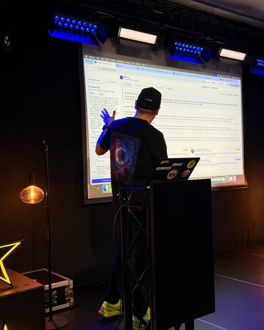A man in a black t-shirt and cap stands on a stage, pointing at a large projection screen displaying a software dashboard during a presentation.