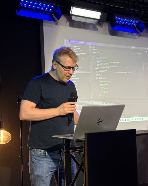 A man stands at a podium, holding a microphone and looking down at a laptop while a screen behind him displays lines of Python code.