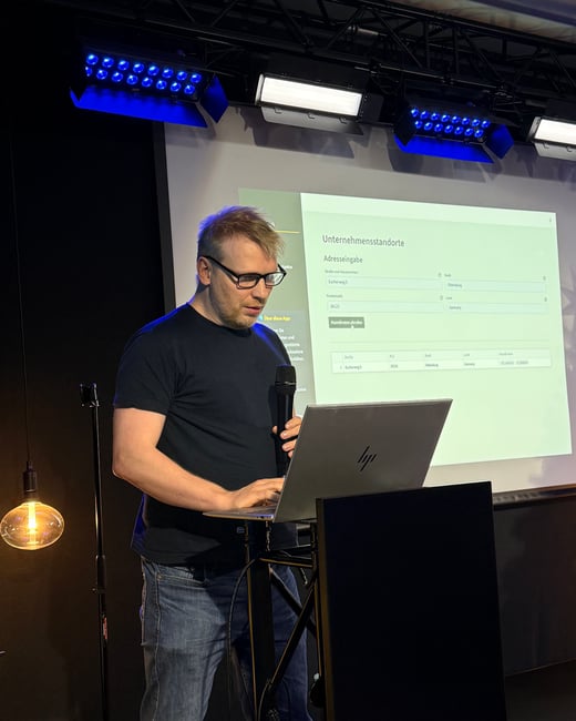 A man stands at a podium, speaking into a microphone while operating a laptop during a presentation. A screen behind him displays a German-language web application for managing company locations.