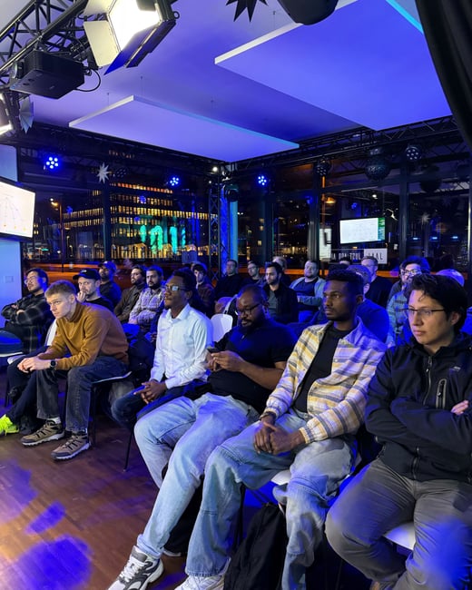 A diverse audience of men is seated in a dimly lit room with blue ambient lighting, attentively watching a presentation shown on screens.