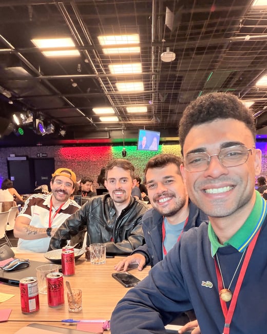 A group of four men smiling for a selfie while sitting at a table in an indoor event space with Coca-Cola cans and laptops.