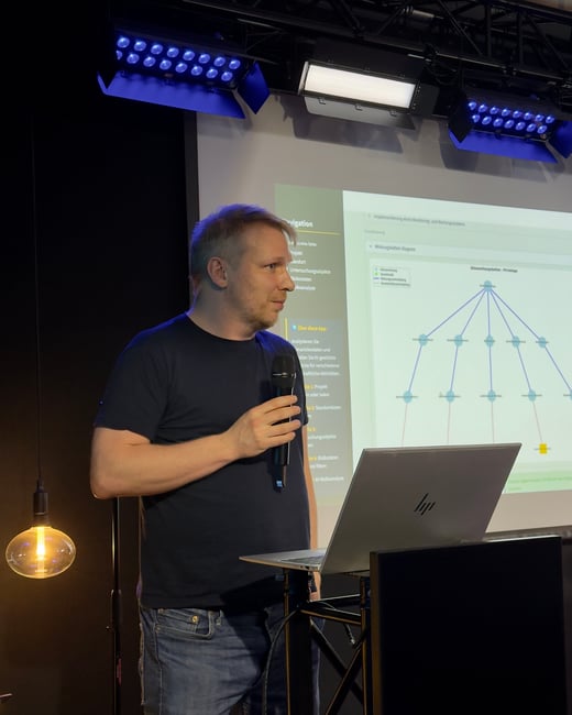 A man in a dark t-shirt and jeans is giving a presentation, holding a microphone while standing next to a laptop with a projection screen in the background.