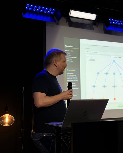 A man stands on a stage holding a microphone, giving a presentation in front of a large screen displaying a technical software interface.