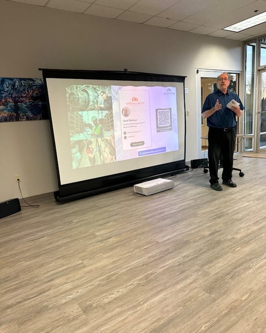 A man is giving a presentation next to a large projection screen in a modern indoor space with light wood-look flooring. The screen displays a digital contact card for "Lloyd Spencer," featuring workplace images and a large QR code.
