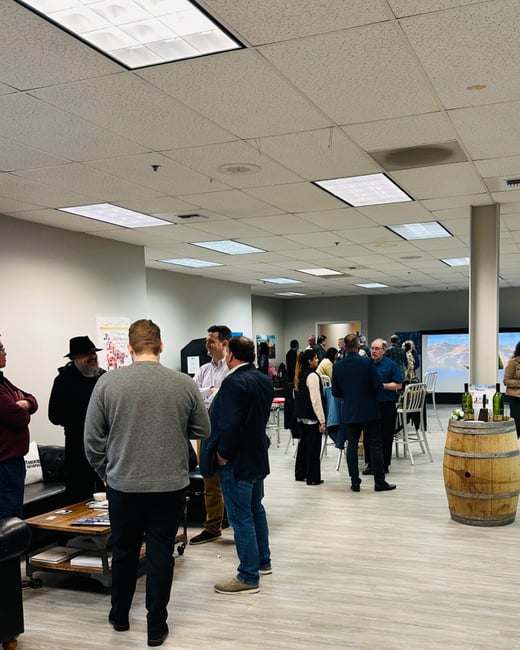 A photograph captures an indoor networking or social event where a large group of people are standing in small clusters, engaged in conversation within a brightly lit room. A wooden barrel topped with wine bottles stands prominently in the foreground, suggesting a wine tasting or reception.