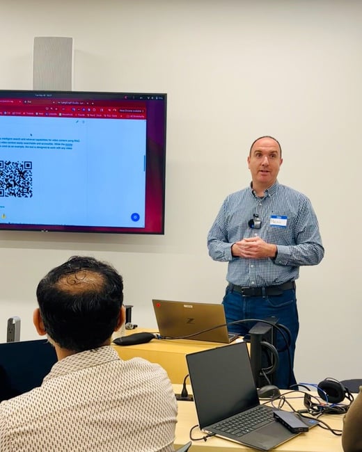 A man wearing a plaid shirt and a name tag is giving a technical presentation in an office setting, standing next to a large screen displaying a QR code and text about intelligent search capabilities. The back of an audience member's head is visible in the foreground, along with several laptops on a table.