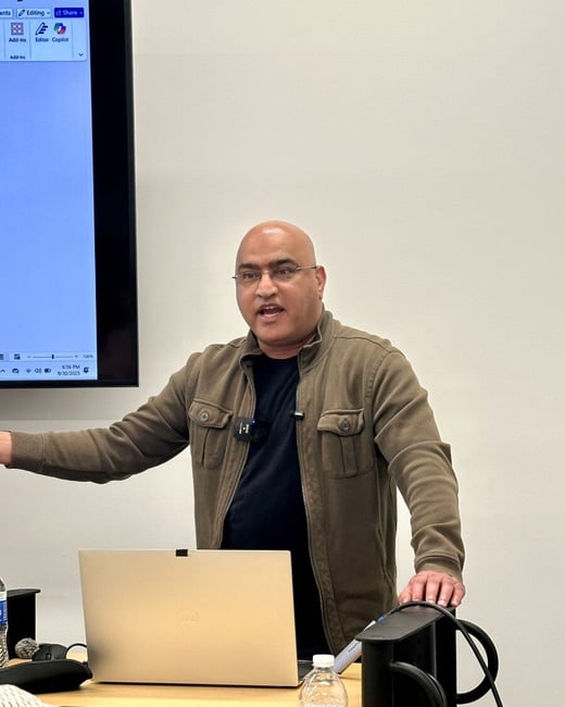 A photograph captures a bald man wearing glasses and a khaki jacket, speaking passionately while standing behind a podium with a laptop and water bottles, likely giving a presentation in a brightly lit indoor venue.