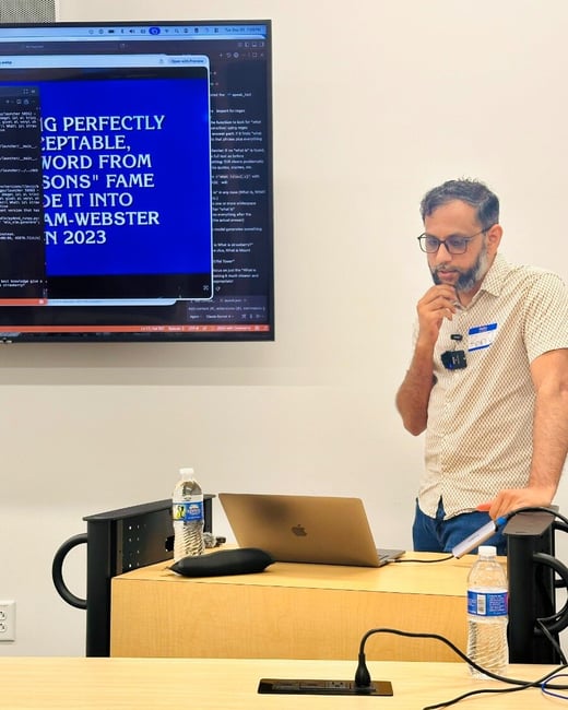 A photograph capturing a man standing next to a wooden podium, giving a technical presentation or demonstration in a conference room setting, with a large screen displaying code and a blue presentation slide.