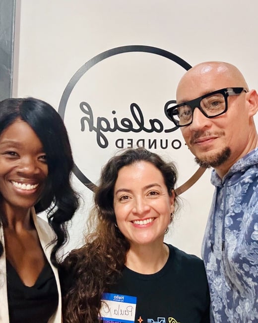 A close-up group photo, likely a selfie, of three smiling people (two women and one man) standing indoors in front of a white wall featuring a large black logo.