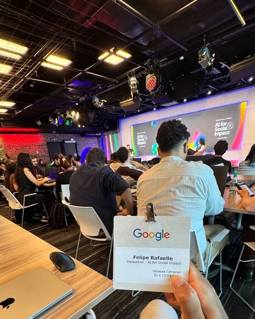 A first-person perspective photo of a person holding a Google event badge for an 'AI for Social Impact' hackathon, with a crowded room and a presentation screen in the background.
