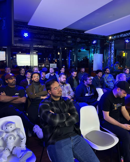 An audience of mostly young men sits in a dimly lit room with blue stage lighting, attentively watching a presentation. A small humanoid NAO robot is seated in a white chair in the front row.