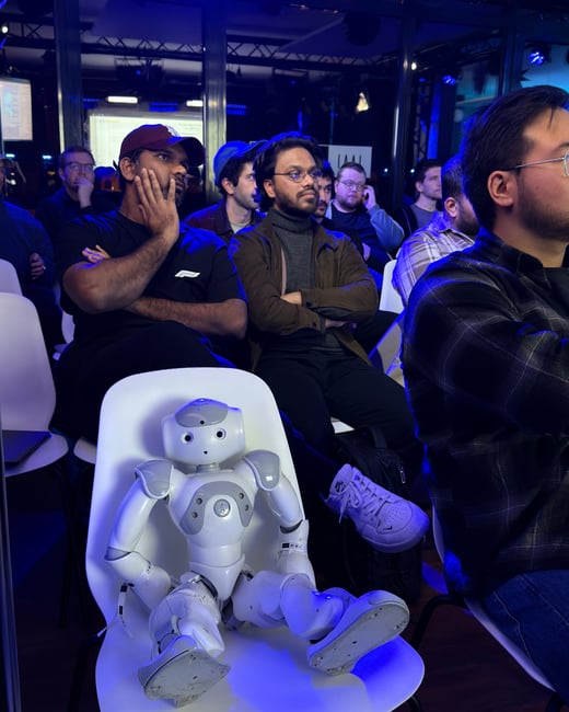A small white humanoid robot sits in a white chair among an audience of people at an indoor event, appearing to attend a presentation.