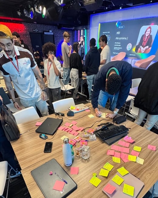 A group of people are gathered around a large wooden table covered in laptops and numerous colorful sticky notes during a workshop or hackathon. In the background, a large screen displays a presentation slide with a woman's photo and contact information.