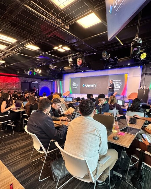 A photograph of a tech event or workshop titled 'AI for Social Impact' featuring Google's Gemini, with attendees working on laptops while a presenter stands on stage.