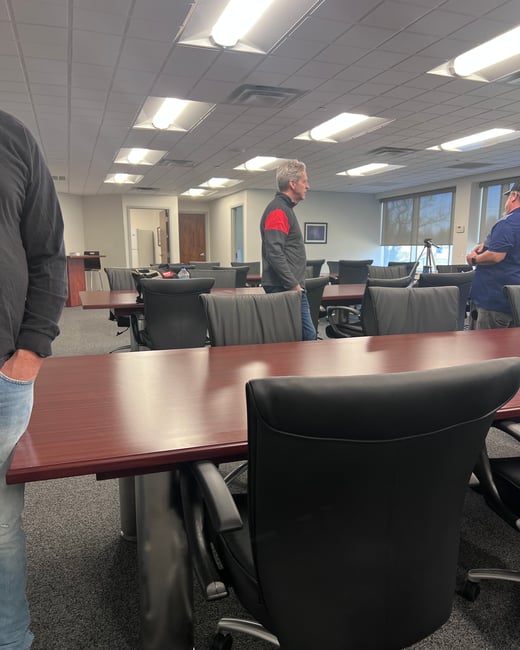 An indoor view of a conference or training room with long brown tables and black chairs, where three men are standing.