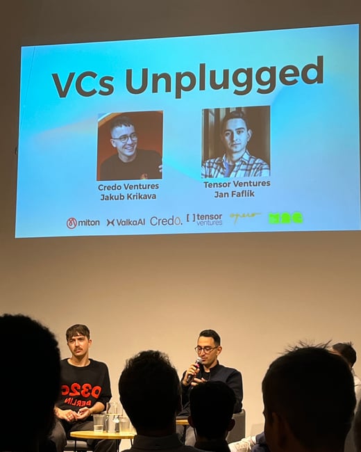 A photograph capturing an indoor panel discussion or presentation titled "VCs Unplugged," showing two male speakers seated on a stage while a large projection screen behind them displays their names, firms, and sponsor logos.