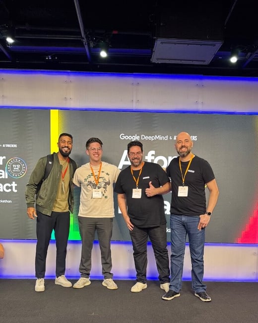 Four men pose for a photo on a stage in front of a large digital screen displaying event information for a hackathon.