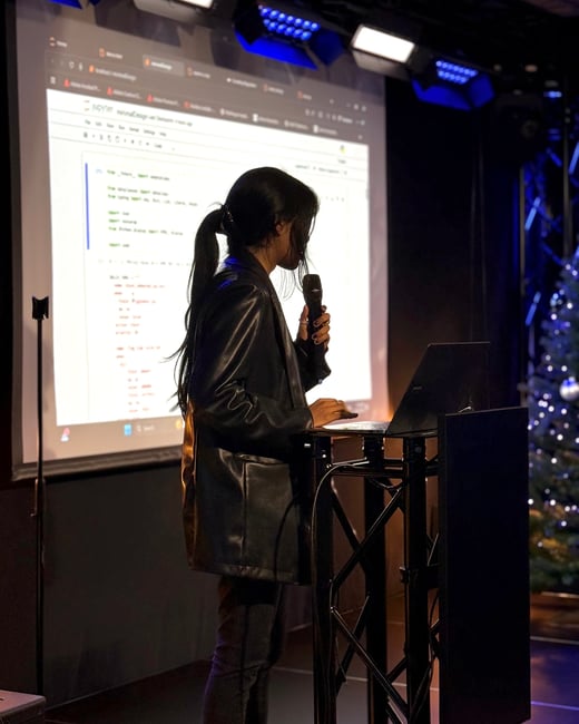 A woman stands at a podium giving a presentation, holding a microphone and using a laptop while a Jupyter notebook is projected on the screen behind her.