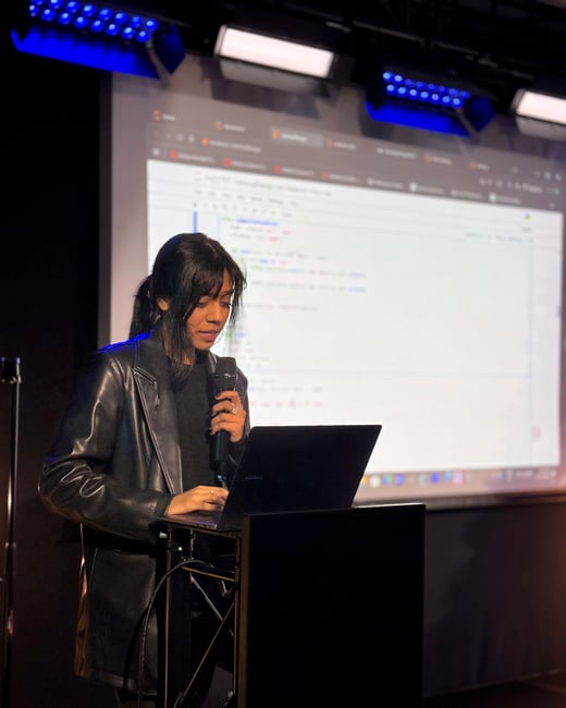 A woman in a black leather jacket is giving a presentation, holding a microphone and looking down at a laptop while a screen behind her displays code.
