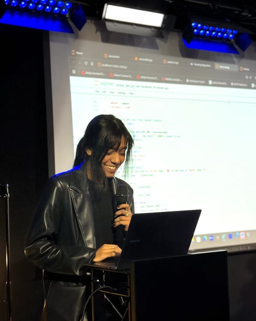 A young woman in a black leather jacket is giving a technical presentation, holding a microphone and using a laptop in front of a large screen displaying Python code in a Jupyter notebook.
