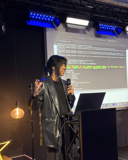 A woman in a black leather jacket is giving a presentation, holding a microphone and gesturing while standing in front of a screen showing code and terminal output.
