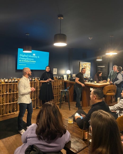 A candid photograph of an indoor professional event or networking session taking place in a dimly lit venue, likely a wine bar, where a man and a woman are presenting near a screen displaying the title 'Digital Identity & Compliance.'