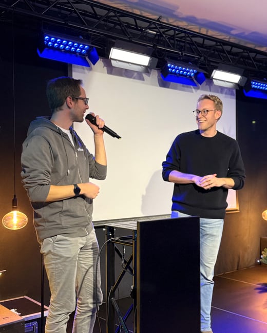 Two men are on a stage during an event; one is speaking into a microphone while the other listens and smiles.