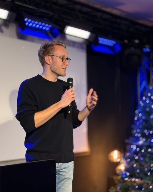 A man wearing glasses and a black sweater is speaking into a microphone on a stage, with a decorated Christmas tree visible in the background.