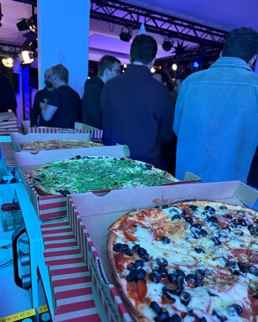 A row of open pizza boxes with various toppings sits on a table in a room with strong blue lighting, with several people socializing in the background.