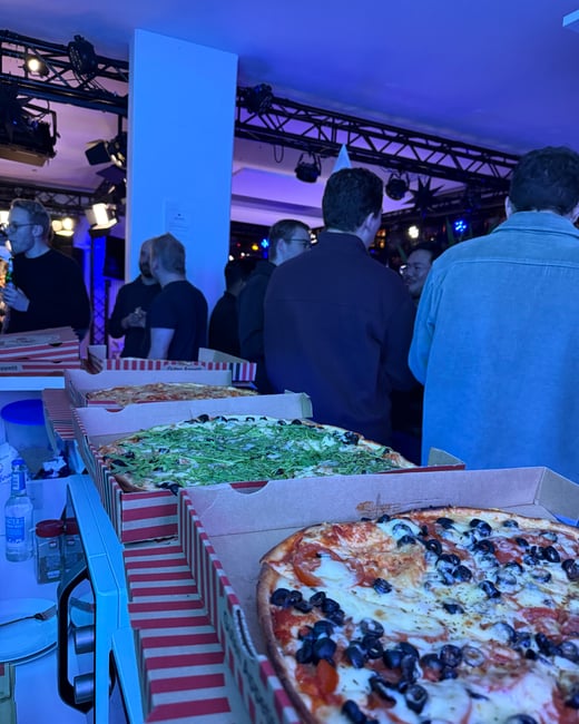 A social gathering in an indoor space featuring several open pizza boxes in the foreground and a group of people socializing under blue ambient lighting in the background.