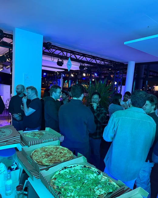 A group of people are gathered in a room with strong blue lighting, standing around a table with several open pizza boxes during a social event.