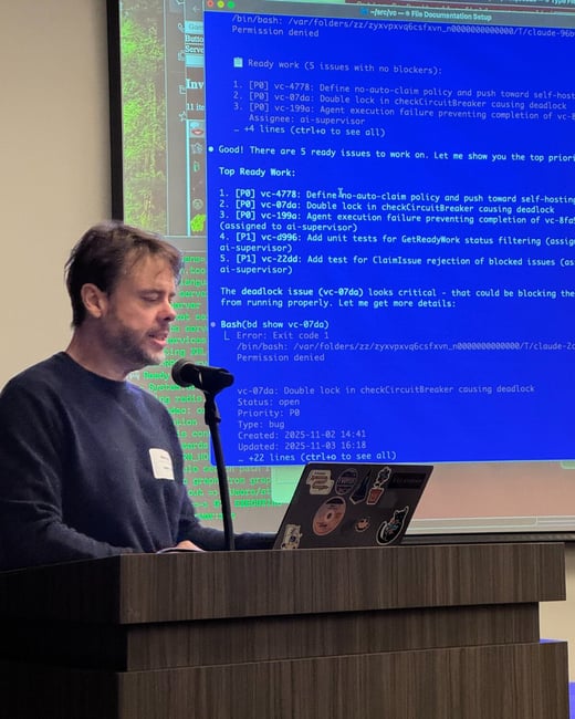 A man wearing a dark sweater is giving a technical presentation from behind a wooden lectern, with a large screen displaying terminal output detailing software issues and status updates behind him.