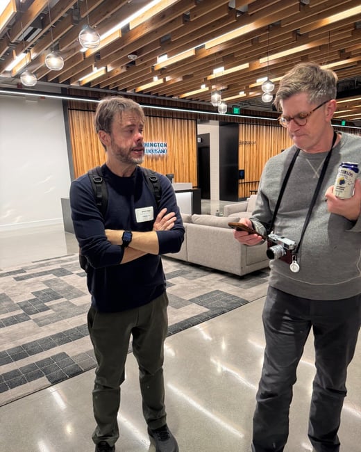 Two middle-aged men are standing and engaged in conversation in a modern indoor lobby or event space characterized by wood paneling and a suspended slat ceiling.