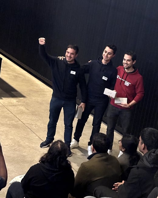 A photograph captures three men standing and celebrating, possibly after receiving awards, while an audience sits on the floor watching them in an indoor event space.