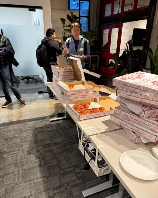 A candid photograph capturing an indoor social gathering, likely in an office setting, where several people are gathered around a long table stacked with numerous pizza boxes, some open and containing pepperoni pizza.