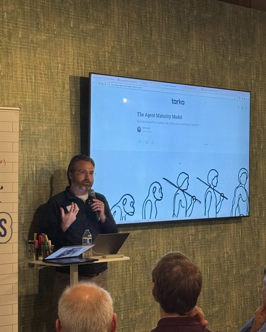 A man is giving a presentation indoors, standing next to a large monitor displaying a slide titled "The Agent Maturity Model" featuring an illustration of human evolution. Several audience members are visible in the foreground, seen from behind.