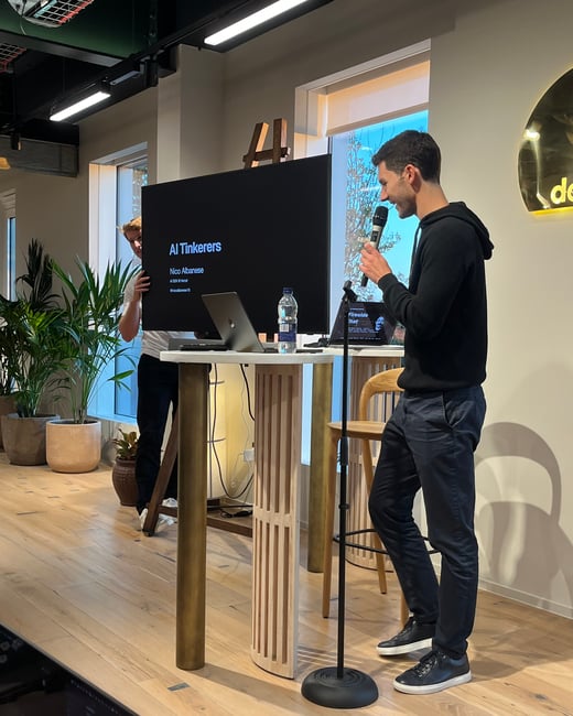 A photograph showing a man presenting at an indoor event, holding a microphone while standing next to a high table with a laptop and a large screen displaying the title 'AI Tinkerers'. Another person is partially visible adjusting the screen setup in the background.