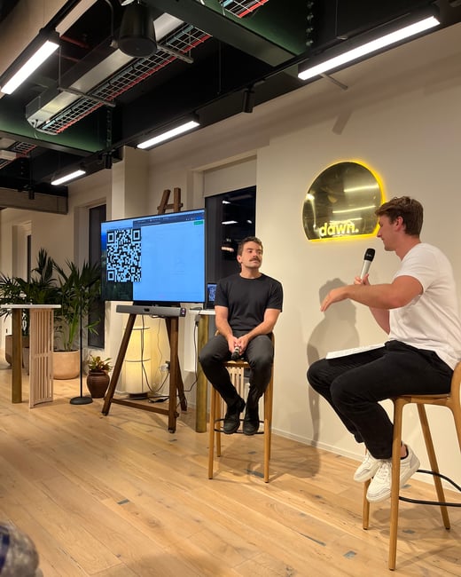 A photograph capturing an indoor event, likely a fireside chat or interview, featuring two men seated on high stools in front of a large screen displaying a QR code and a backlit wall sign reading "dawn."