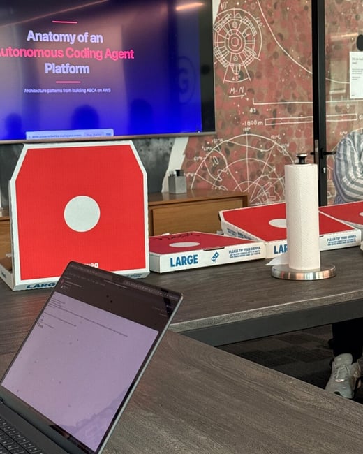 A photograph of an office meeting room featuring several pizza boxes on a table, a laptop in the foreground, and a large screen displaying a presentation about autonomous coding agents.