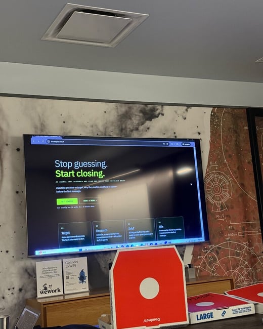 A photograph showing a large television screen in an office or coworking space displaying a website for 'Zulu', with Domino's pizza boxes on a table in the foreground.