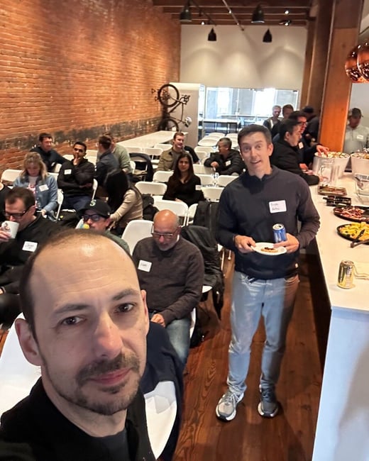 A group of people are gathered in an indoor event space with exposed brick walls and wooden floors, some sitting in chairs while others stand near a food counter.