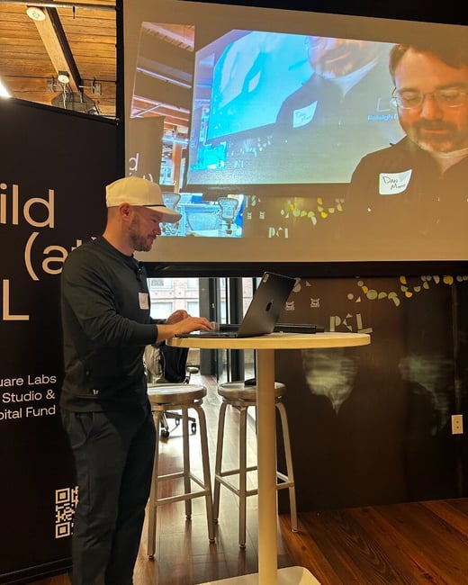 A man in a white baseball cap stands at a high table using a laptop, with a large screen behind him displaying a video call of another person.