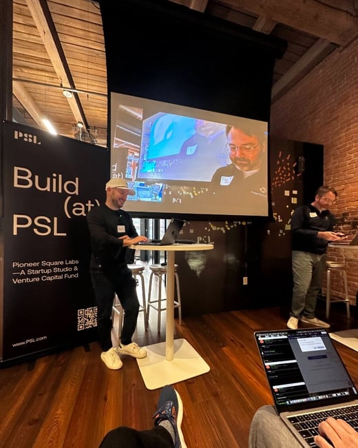 A man in a white cap stands at a high table with a laptop during a presentation at Pioneer Square Labs, with a remote participant visible on a large screen behind him.