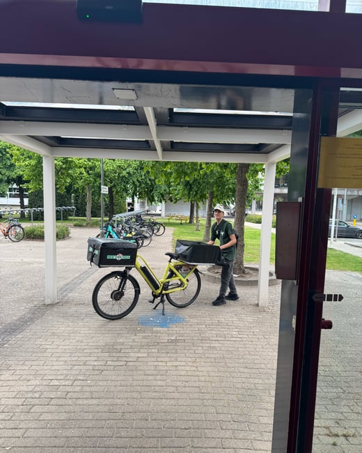 A delivery person stands next to a yellow electric bike equipped with 'New York Pizza' delivery bags under a white outdoor shelter.