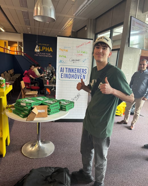 A young man in a green t-shirt and 'NEW YORK' cap gives two thumbs up next to a table with pizza boxes and an 'AI TINKERERS EINDHOVEN' banner.