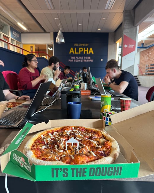 A group of people are sitting at a long table in a coworking space called ALPHA, eating pizza while working on their laptops.