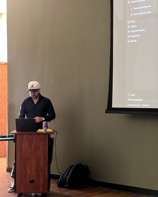 A man wearing a baseball cap stands at a wooden podium with a laptop, giving a presentation next to a large screen displaying a software interface.